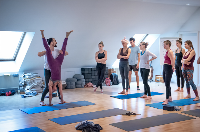 Alexander Kröker zeugt Handstand mit Yogaschülerin mit Gruppe Yogaschülerinnen und Yogaschülern im großen Raum im Yogastudio Berlin
