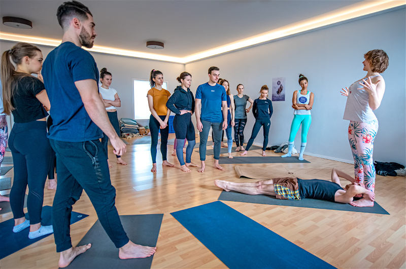 Yogalehrer Weiterbildung Almuth Kramer vor Gruppe zeigt Acroyoga an Yogaschüler, Personen im Hintergrund lächeln, Events Workshop
