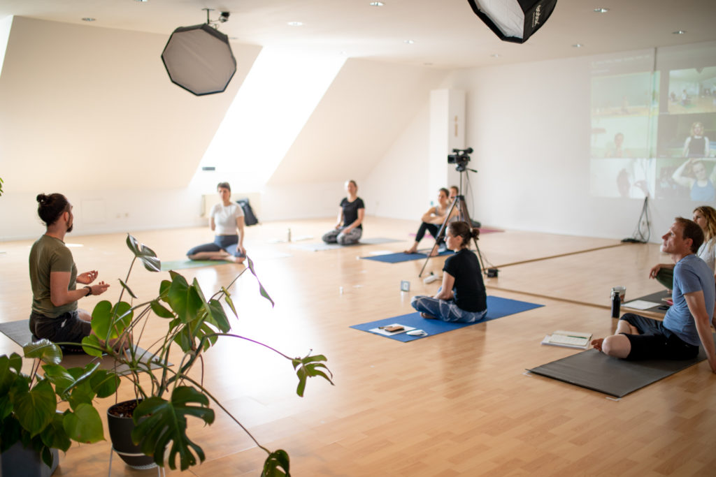 Alexander Kröker im großen Raum sitzt auf Yogamatte und erklärt Yogaschülerinnen und Yogaschülern im Yogastudio, eine Kamera ist im Rau und im Hintergrund Zoom an der Wand bei einer Ausbildung Berlin
